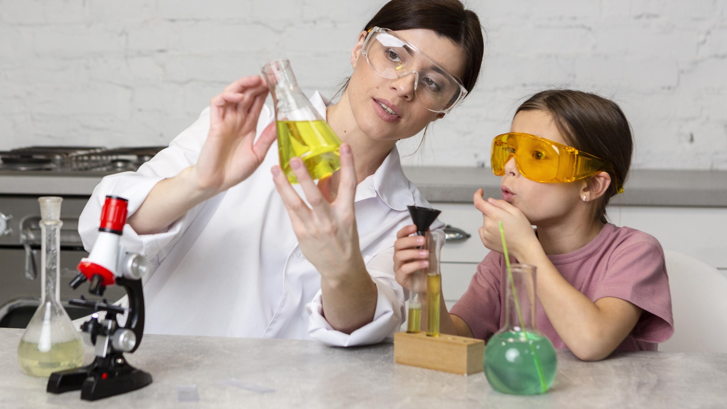 A woman and a girl conducting a science experiment in a laboratory. The woman holds a beaker with yellow liquid, while the girl, wearing safety goggles, watches with curiosity. There are various laboratory equipment, including test tubes, a microscope, and flasks, on the table.