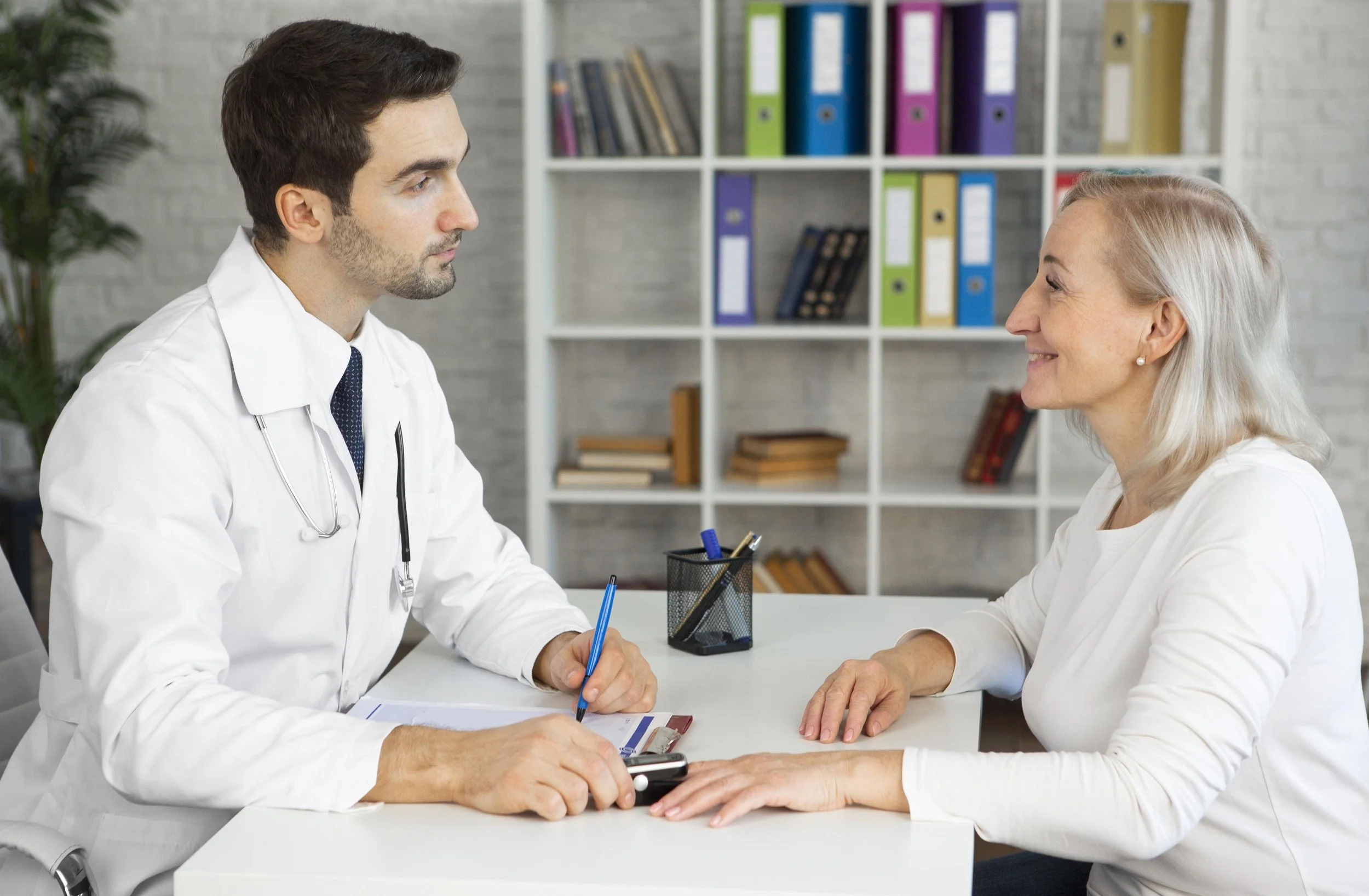 A male doctor in a white coat and a female patient with gray hair are sitting across from each other at a desk, having a consultation in an office with a bookshelf in the background.