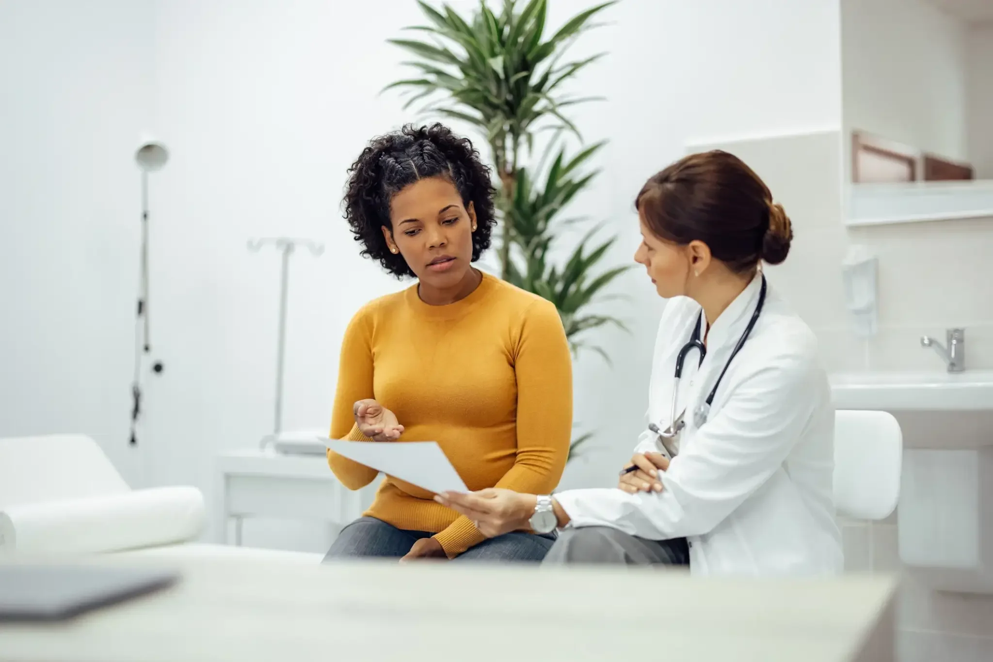 A doctor and a patient having a discussion in a hospital room, with the patient showing her hand and the doctor holding a medical chart.