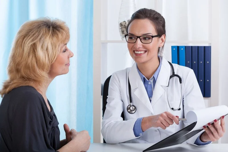 A female doctor with glasses, wearing a white coat and stethoscope, talking to an older woman in a clinical setting.