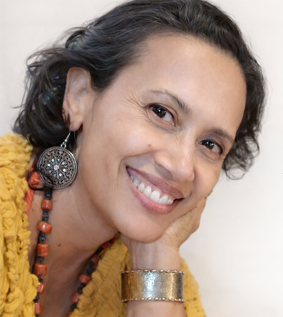 A woman with short dark hair smiling, wearing a yellow textured top, large silver earrings, a chunky gold bracelet, and a red beaded necklace, resting her face on her hand.