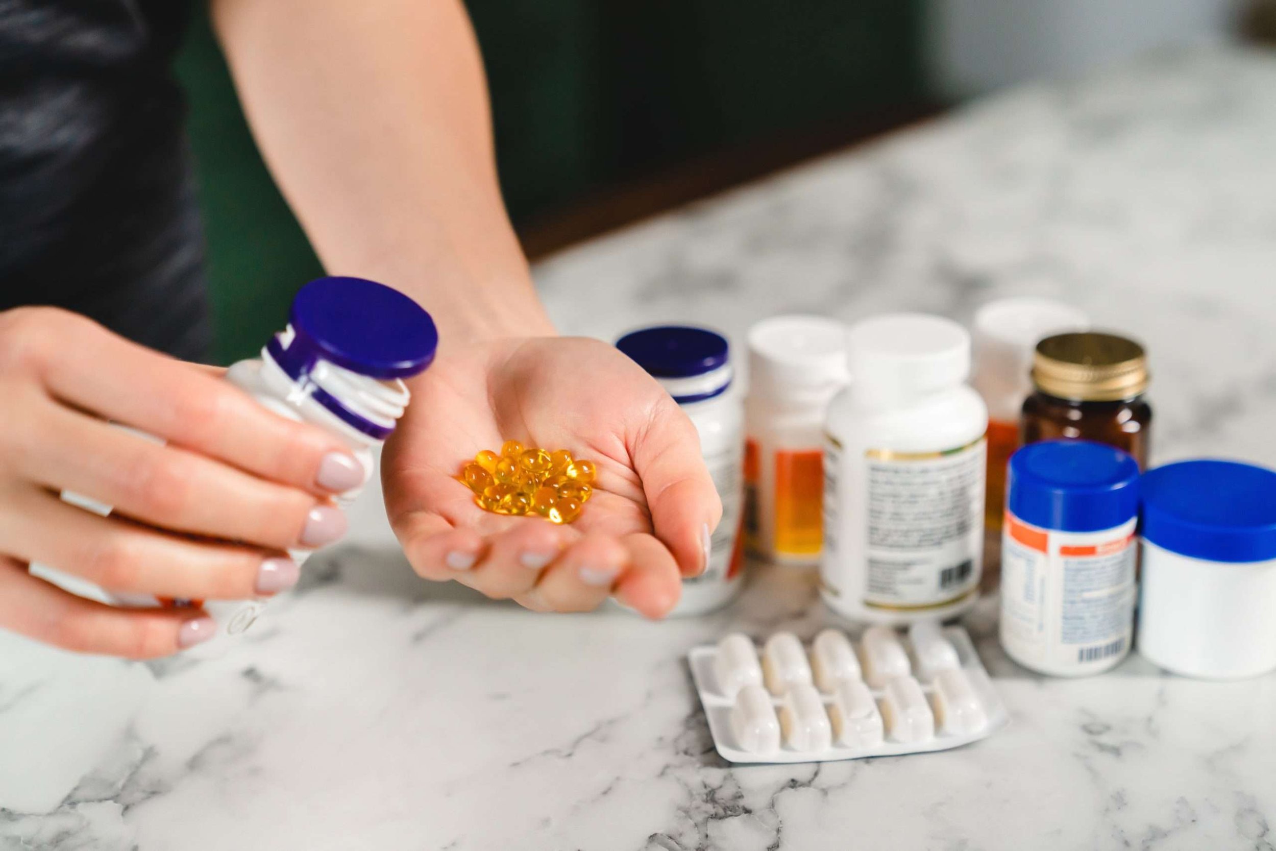Person holding yellow gel capsules and a pill bottle while pouring more capsules into their hand, with various medication bottles on a marble countertop.
