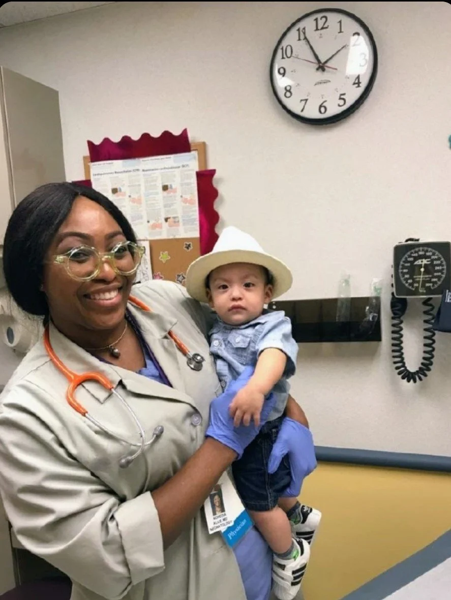 A healthcare professional holding a young boy in a hospital room with a clock showing 1:09 and medical equipment in the background.