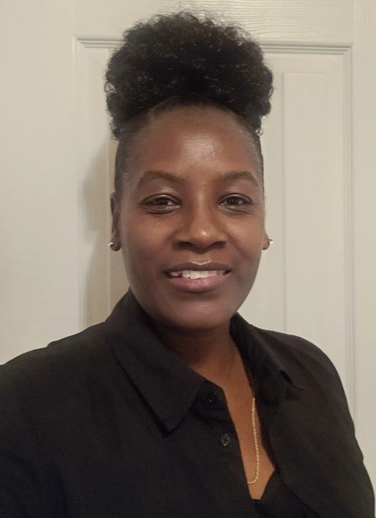 A woman with dark skin and short natural hair tied up in a puff, wearing earrings, a black shirt, and a gold necklace, standing in front of a white door, smiling at the camera.