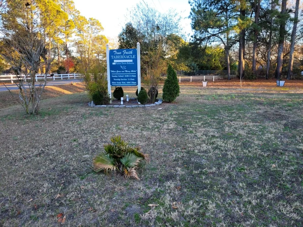 A church sign in a grassy yard surrounded by trees, showing times for Sunday school, worship service, and Bible study.