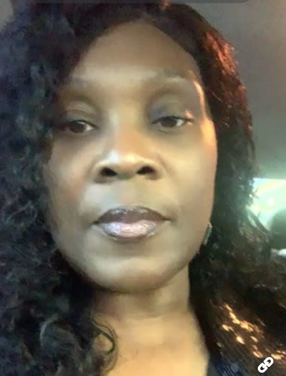 Close-up of a woman with dark, curly hair, wearing makeup including dark lipstick, in a dimly-lit setting.