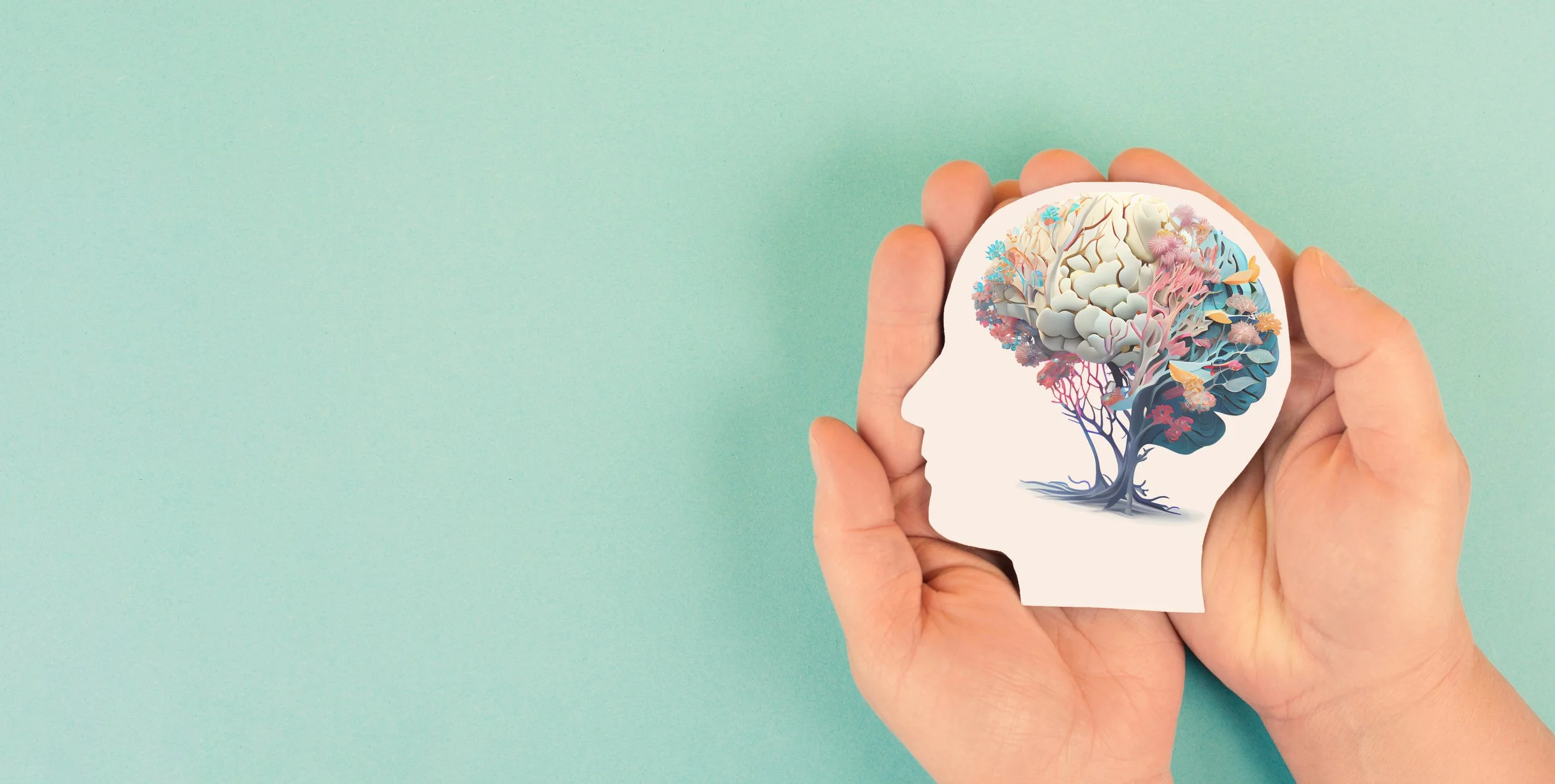 A hand holding a paper cut-out of a human head silhouette with a colorful, artistic illustration of a tree and brain inside. The background is light turquoise.