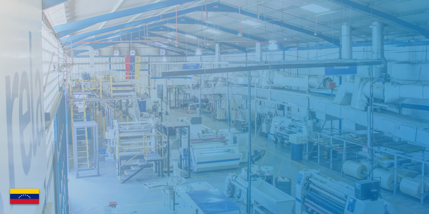 Interior of a large industrial manufacturing plant with machinery, conveyor belts, and equipment, under a curved metal roof with glass panels, with Venezuelan flag in the bottom left corner.
