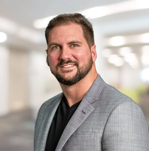 Professional man wearing a gray suit jacket and black shirt, smiling in an indoor setting with blurred background.