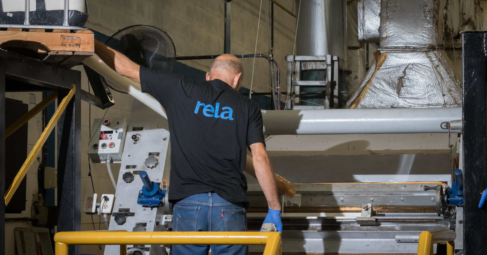 A worker with a black shirt with blue lettering 'rela' on the back operates industrial machinery in a factory.