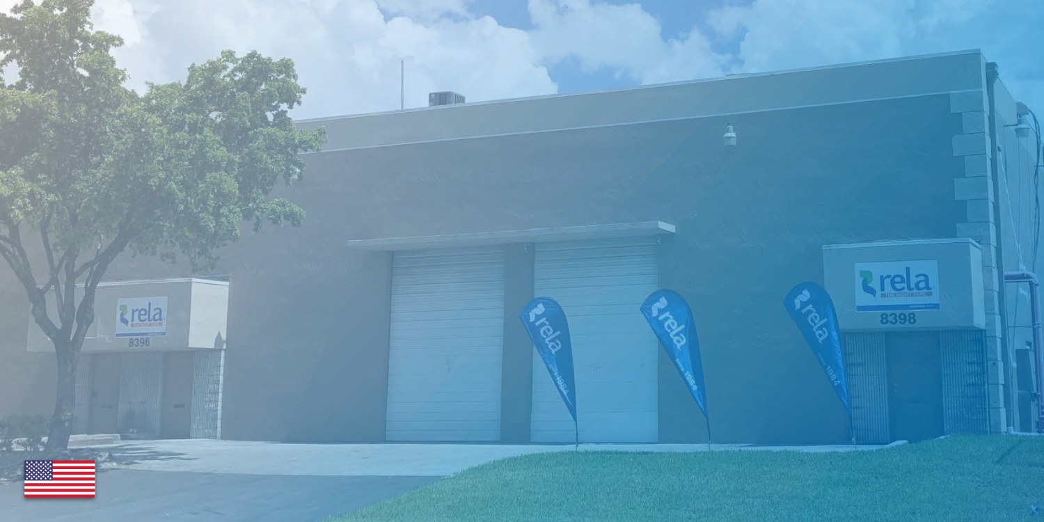 Industrial building with three blue flags bearing the 'rela' logo, and two 'rela' signs on the building, with a tree in the left foreground and a grassy area in the front.