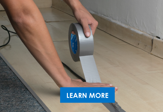 A person's arm applying gray tape to a wooden floor, with a blue 'Learn More' button overlay.