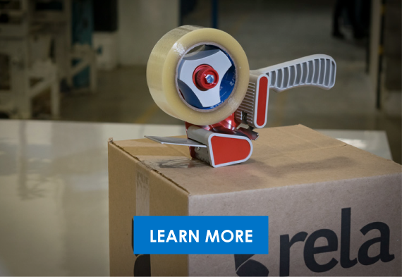 A tape dispenser with a large roll of packing tape mounted on a cardboard box in a warehouse setting.