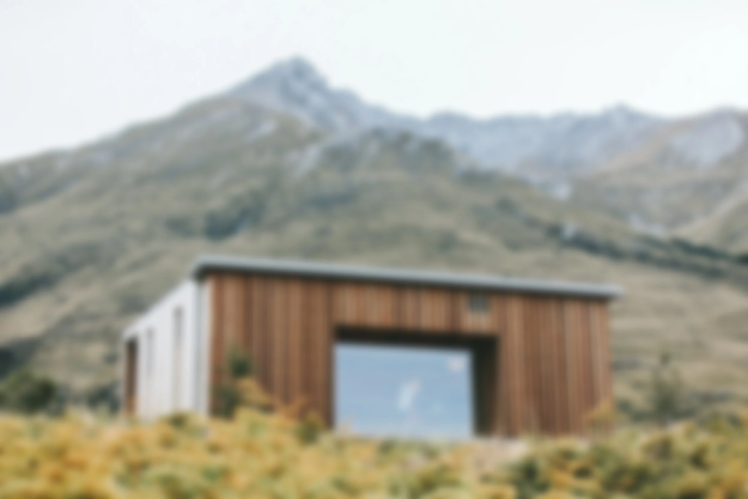 Modern wooden house with large glass window set against a mountainous landscape with dry grass and small trees.