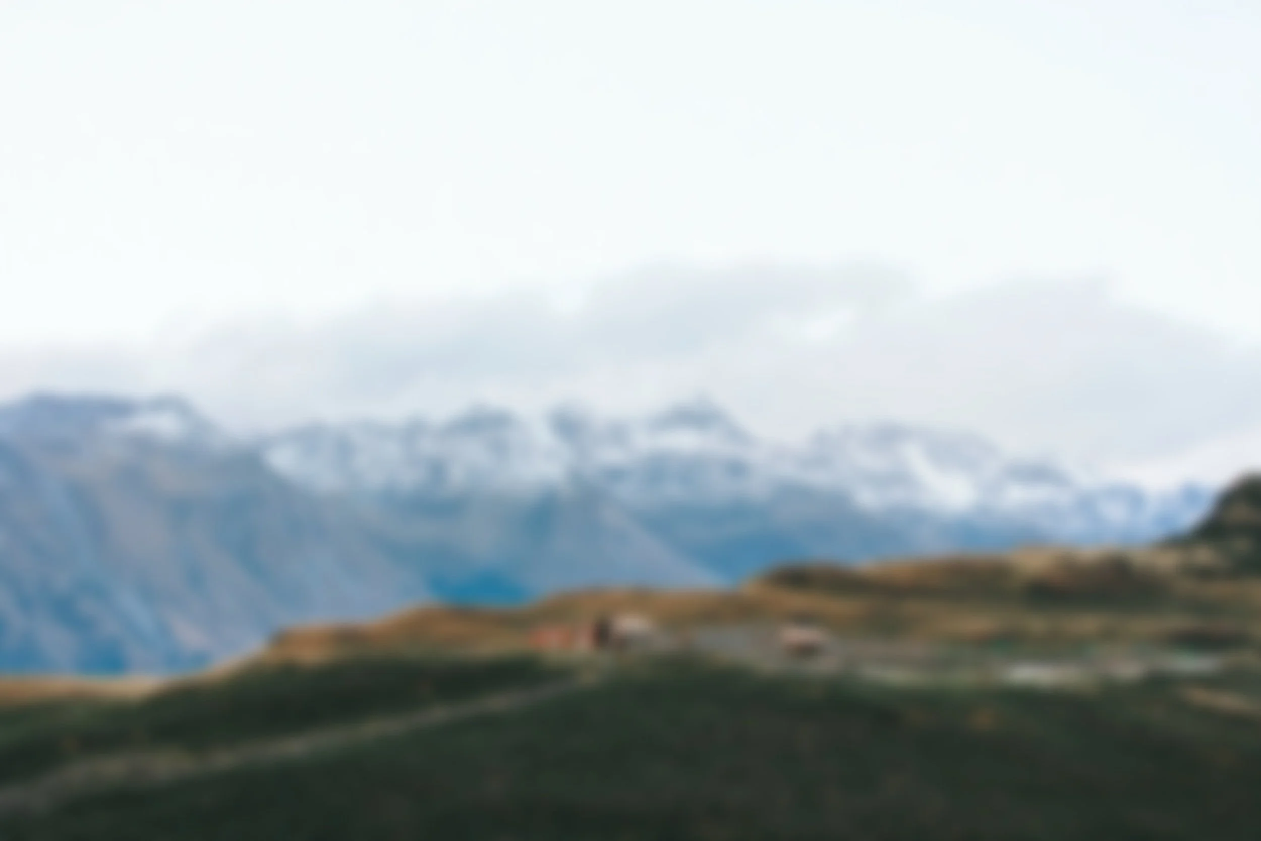 A landscape of rolling hills with a house, mountains with snow, and a cloudy sky in the background.