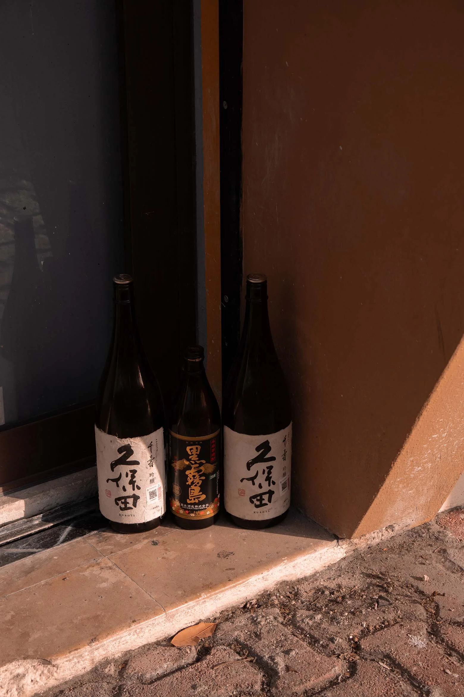 Tres botellas de sake japoneses colocadas en la acera junto a una pared, iluminadas por la luz natural del atardecer que dibuja sombras geométricas.