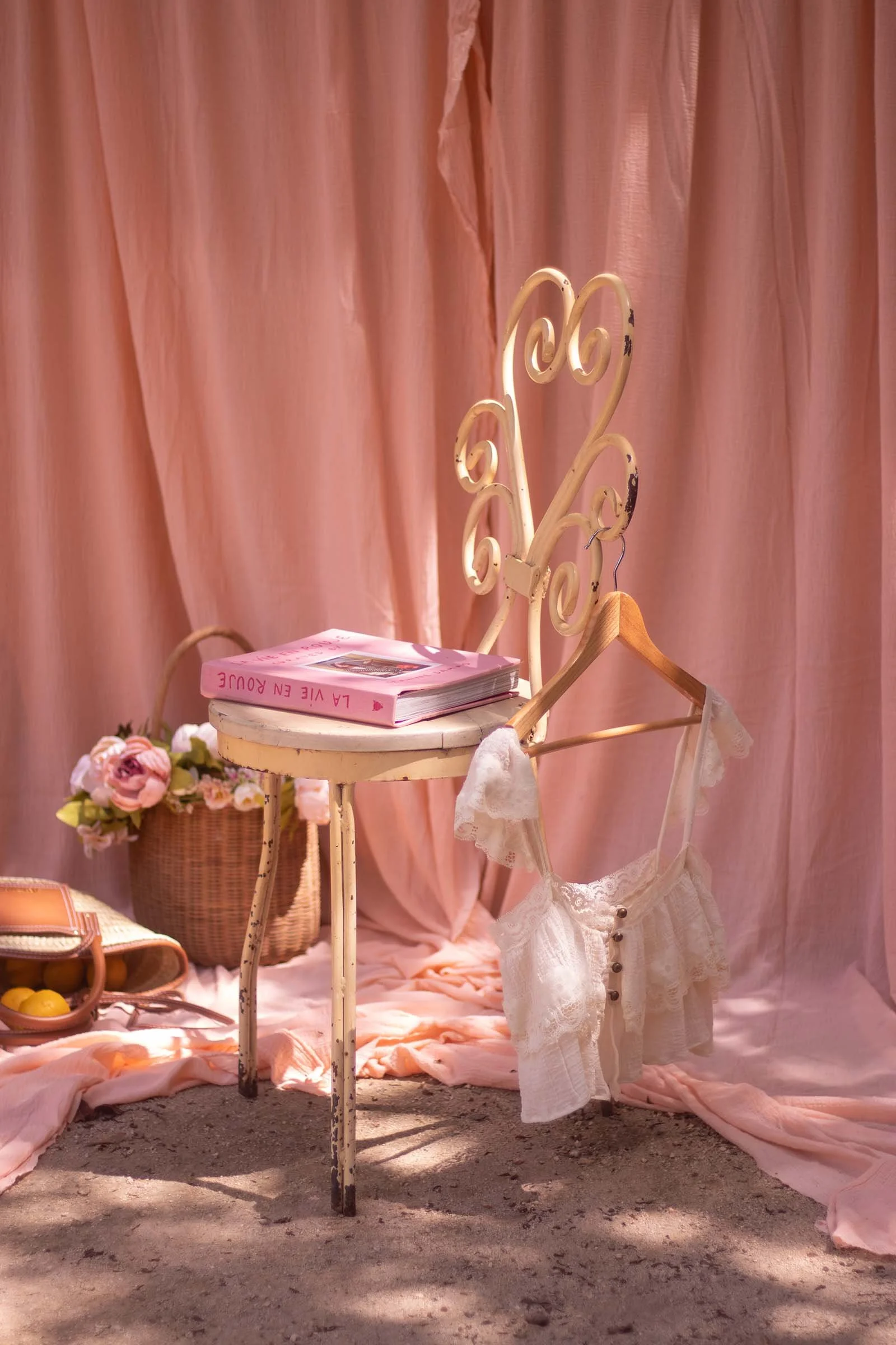 Fotografía de escenario para creación de contenido; ccon cortinas rosas, una silla vintage con un libro rosa, una cesta con flores y un bolso con fruta.