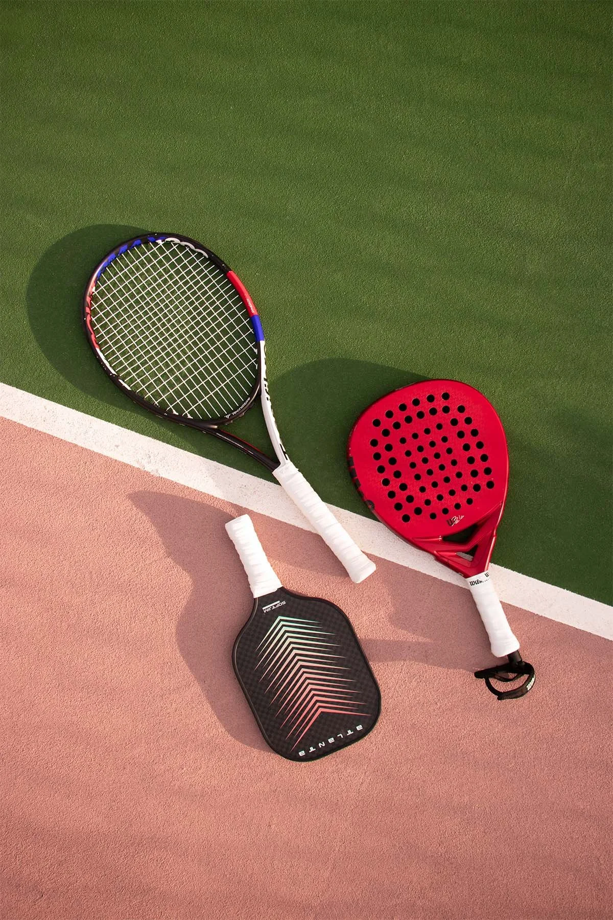 Fotografía editorial deportiva, sobre la cancha se ve una raqueta de tenis, una raqueta. de pádel y una raqueta de pickleball.