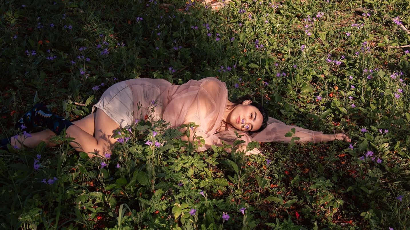 Fotografía editorial de joven mujer acostada en un campo lleno de flores moradas y verdes, con ropa de color rosa y expresión tranquila.
