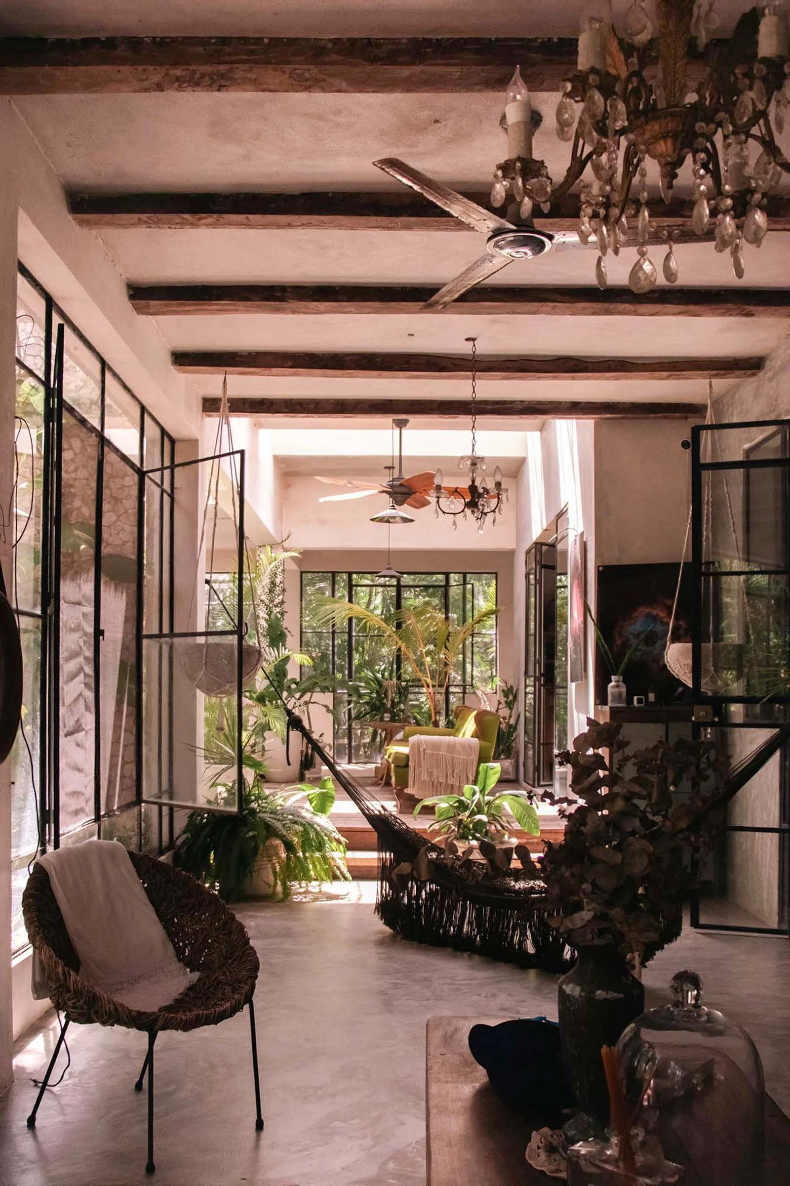 Fotografía de interior de una sala decorada con plantas, sillas de mimbre, mesas, y artículos interesantes, con mucha luz natural y puertas y ventanas grandes.