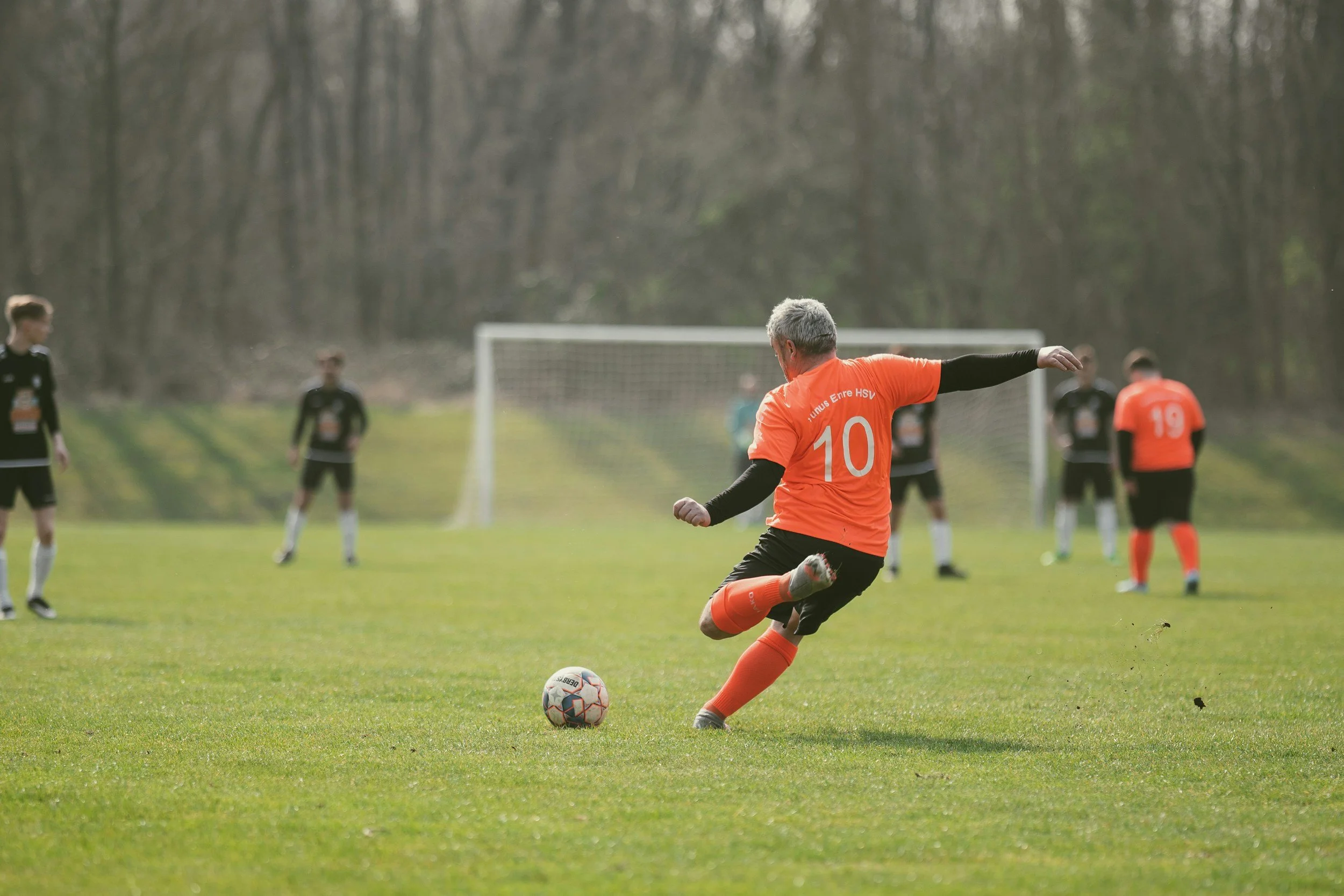 A soccer player in an orange jersey with the number 10 kicks a soccer ball on a grassy field, with other players in black jerseys in the background and a soccer goal.