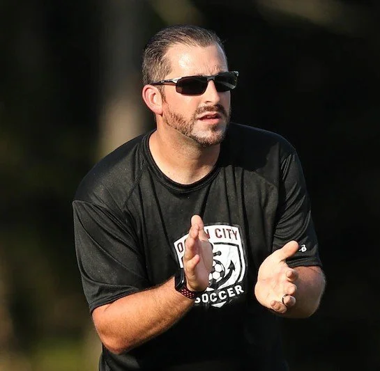 A man wearing sunglasses and a black soccer coach shirt with a logo, standing outdoors, gesturing with his hands.