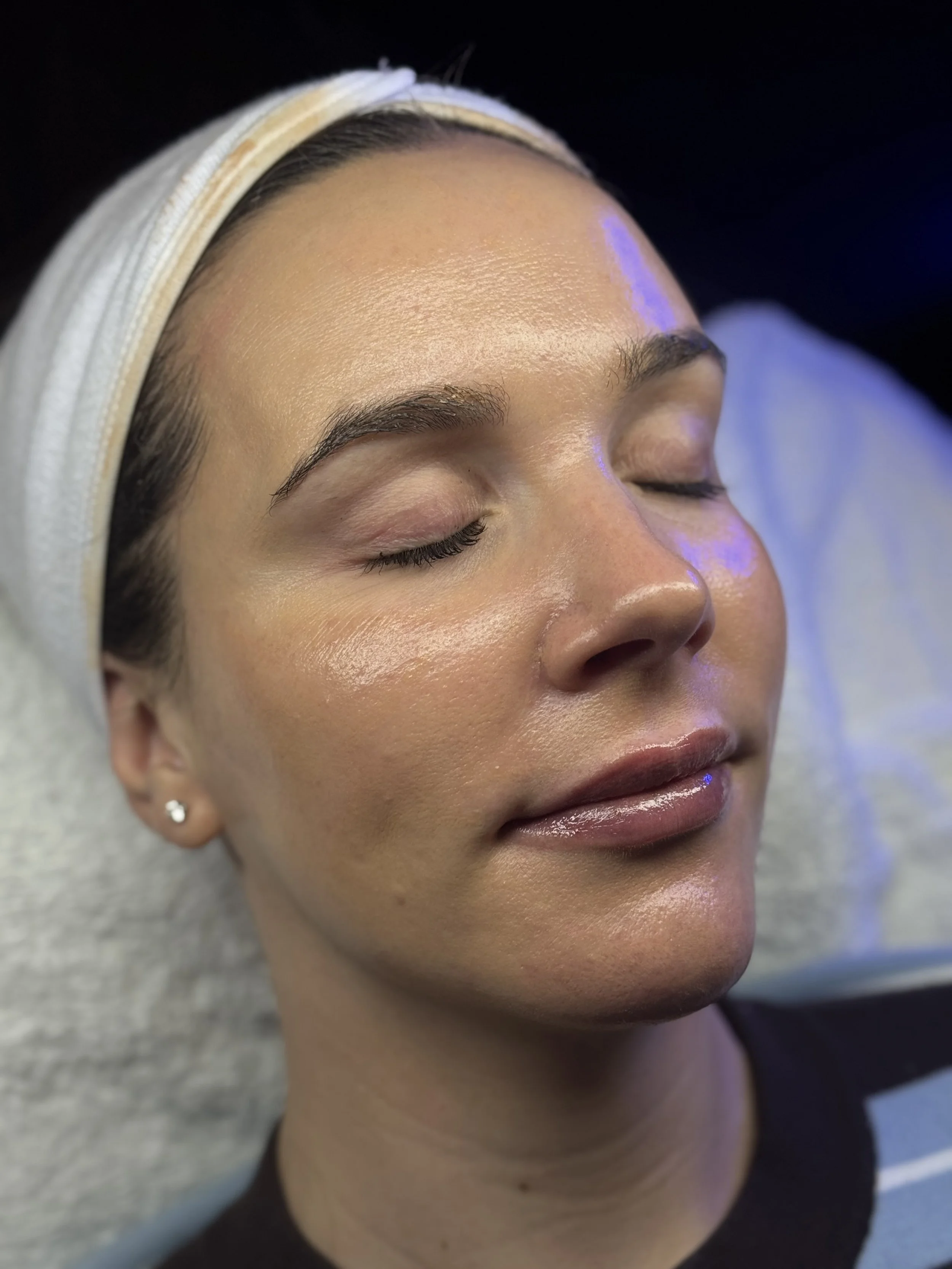 Close-up of a woman's face with eyes closed, showing smooth, glowing skin, well-groomed eyebrows, and shiny lips, with a white headband on her forehead.