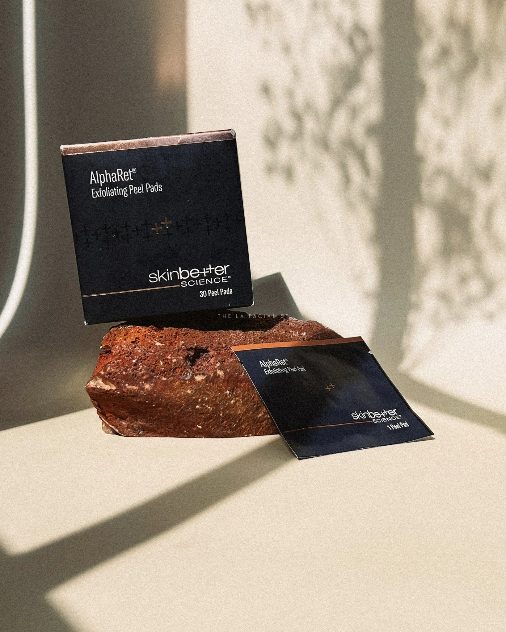Two boxes of Skinbetter Science exfoliating peel pads, one large and one small, placed on a rough stone with shadows cast on a light-colored surface in the background.