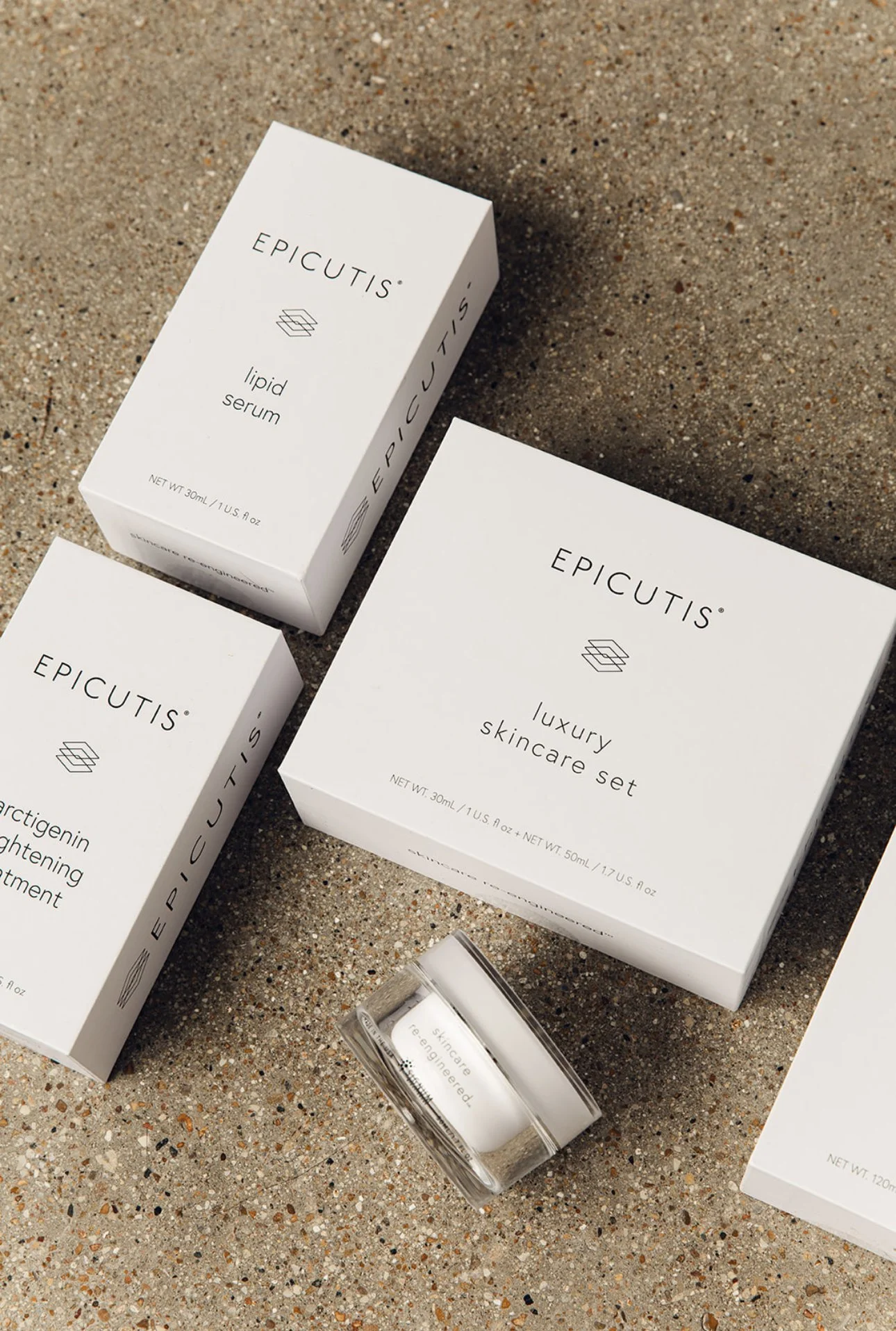 Set of luxury skincare products including a large white box labeled 'luxury skincare set,' a smaller box labeled 'lipid serum,' another smaller box labeled 'arctigenin brightening treatment,' and a small jar with a clear lid, all on a sandy surface.