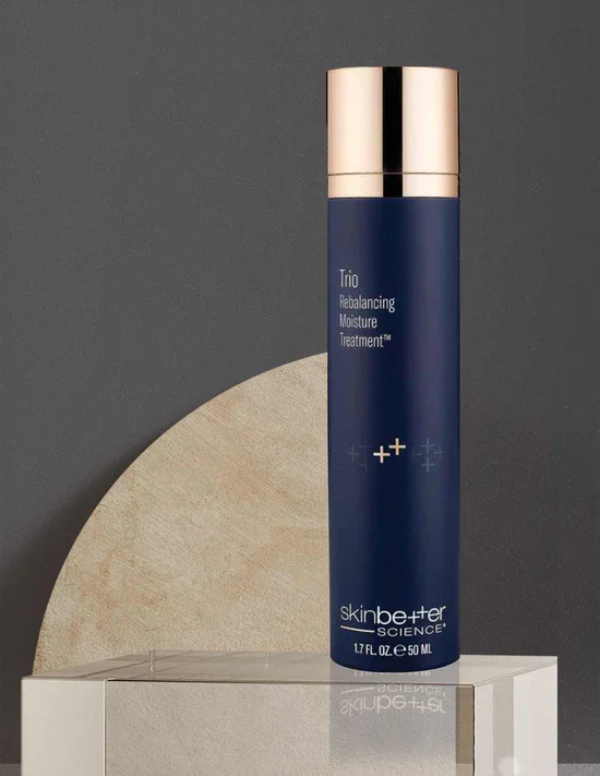 A bottle of skincare product from Skinbetter Science, labeled 'Trio Rebalancing Moisture Treatment,' standing on a white surface with a decorative beige half-moon shape against a dark gray background.