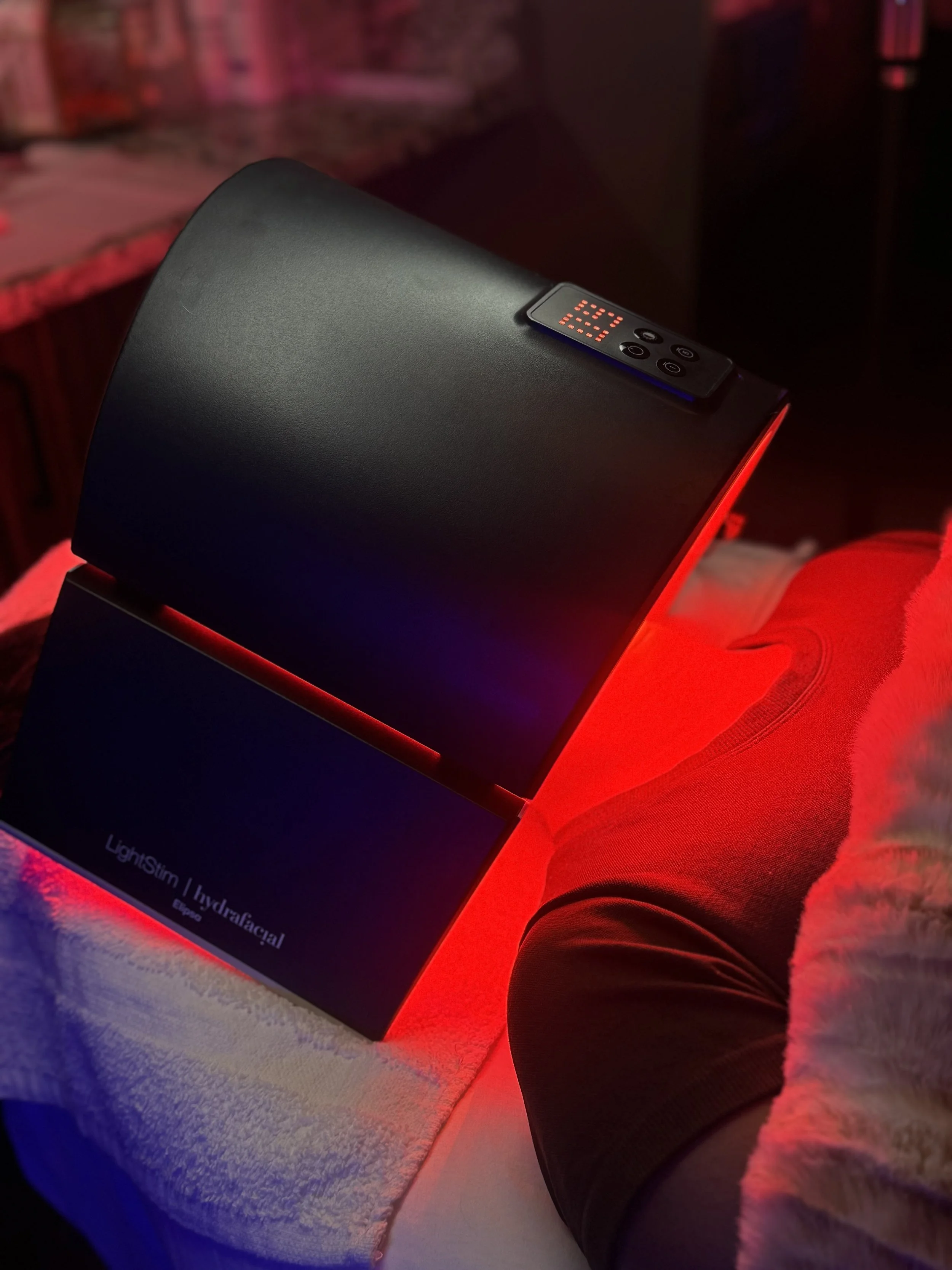 A person lying down with a LightStim LED facial therapy device resting on their lap, glowing red light illuminating the skin. The person is wearing black shorts and a white fuzzy robe, in a room with soft ambient lighting.