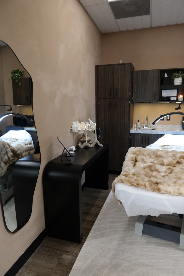 Medical or spa treatment room with a bed covered in a beige faux fur blanket, dark wood cabinets, a sink, and a large mirror on the wall.