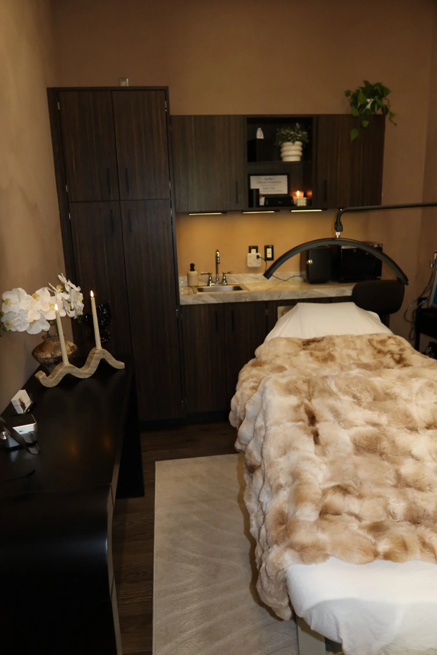 A hospital room with a bed covered in a beige faux fur blanket, a small sink with cabinet, and a black table with a vase of white flowers and candles, featuring warm lighting and wood flooring.