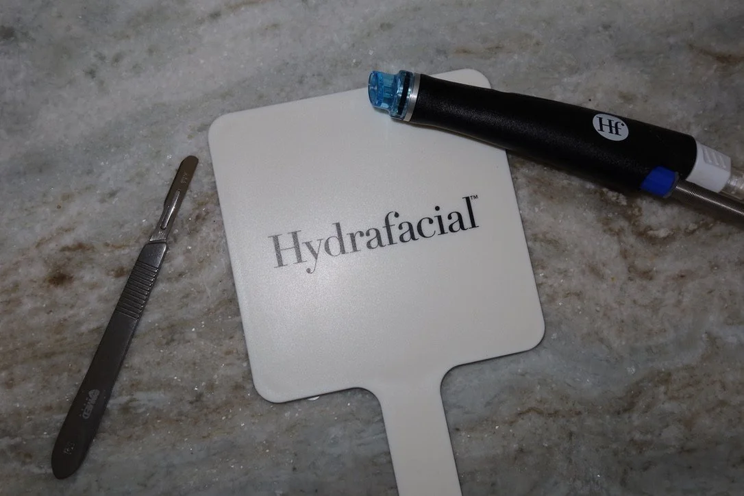 A hydrafacial skin treatment tool, a scraper, and a vacuum device are arranged on a marble surface.