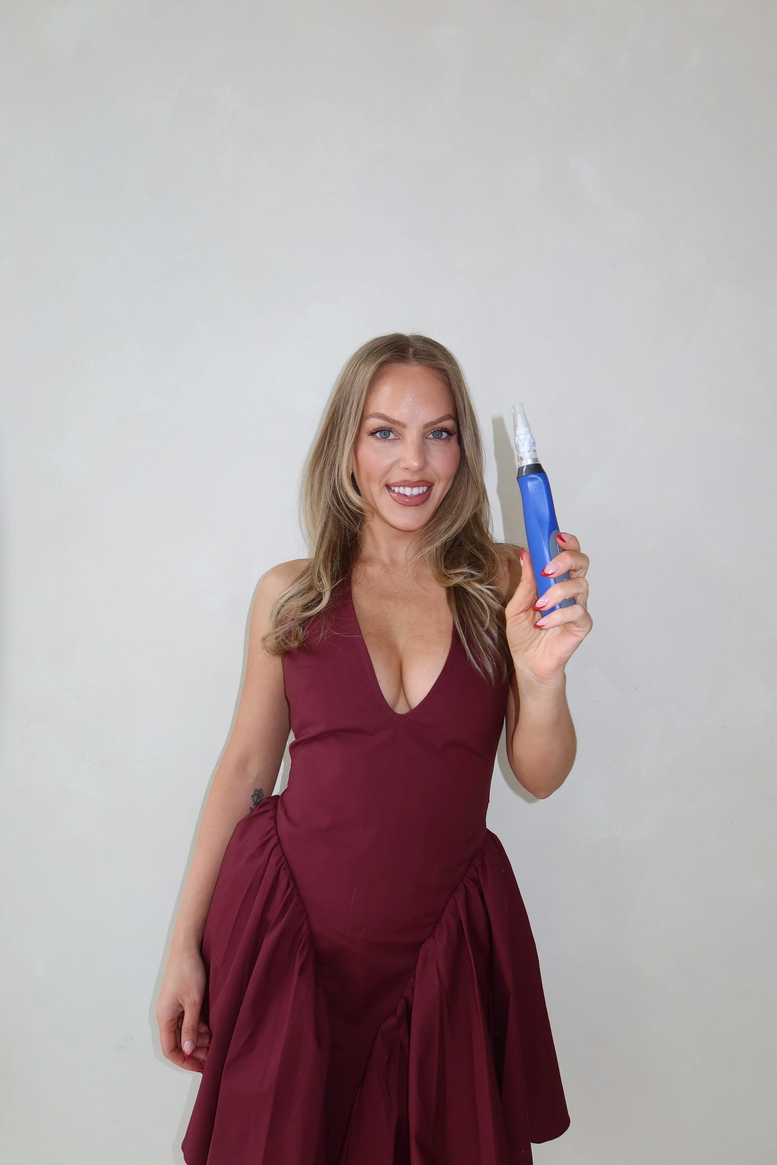 A smiling woman in a burgundy dress holding a blue electric toothbrush against a plain off-white wall.