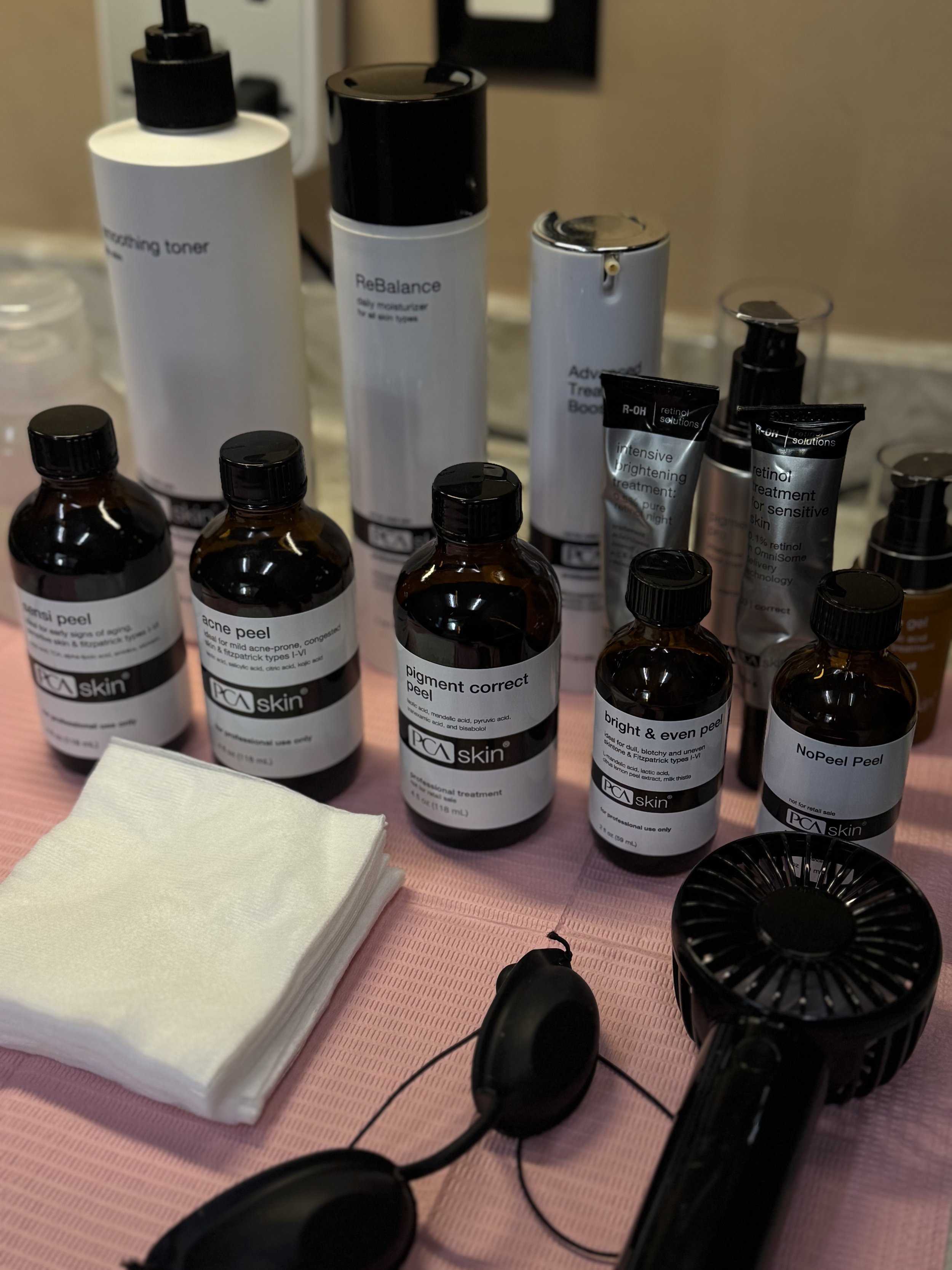 Skincare products including bottles of PCA skin treatments, facial tissues, a fan, and sunglasses on a pink surface.