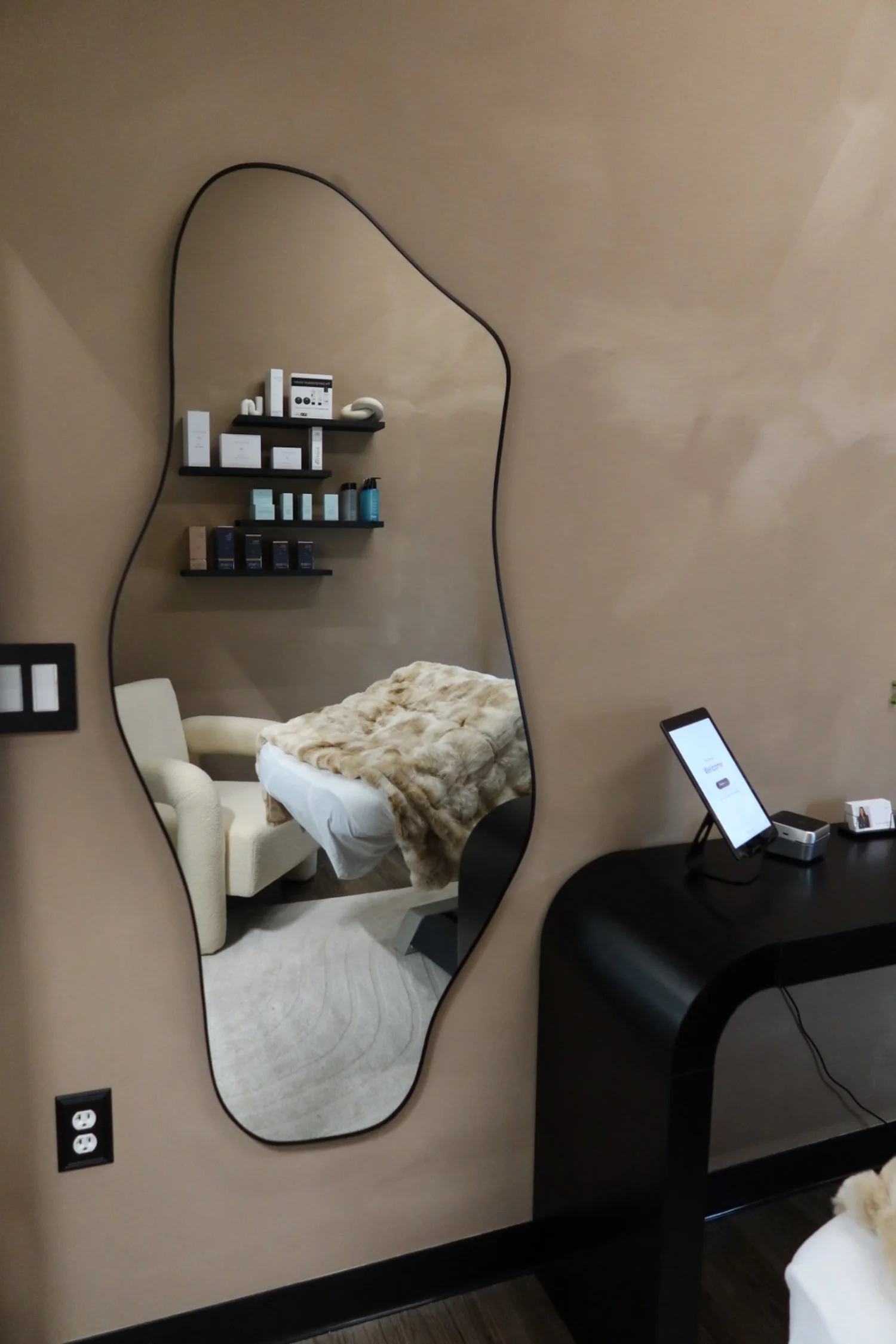 A hotel room mirror with an irregular shape mounted on a beige wall, reflecting a bed with a fur blanket, a white armchair, a set of shelves with toiletries, and part of a black dresser with a tablet.