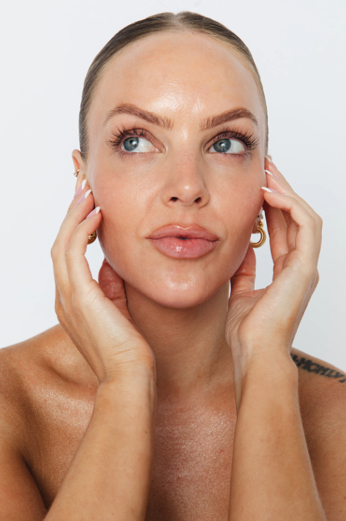 Close-up of a woman with clear skin, light makeup, and gold hoop earrings, holding her face gently with both hands, looking to the side.