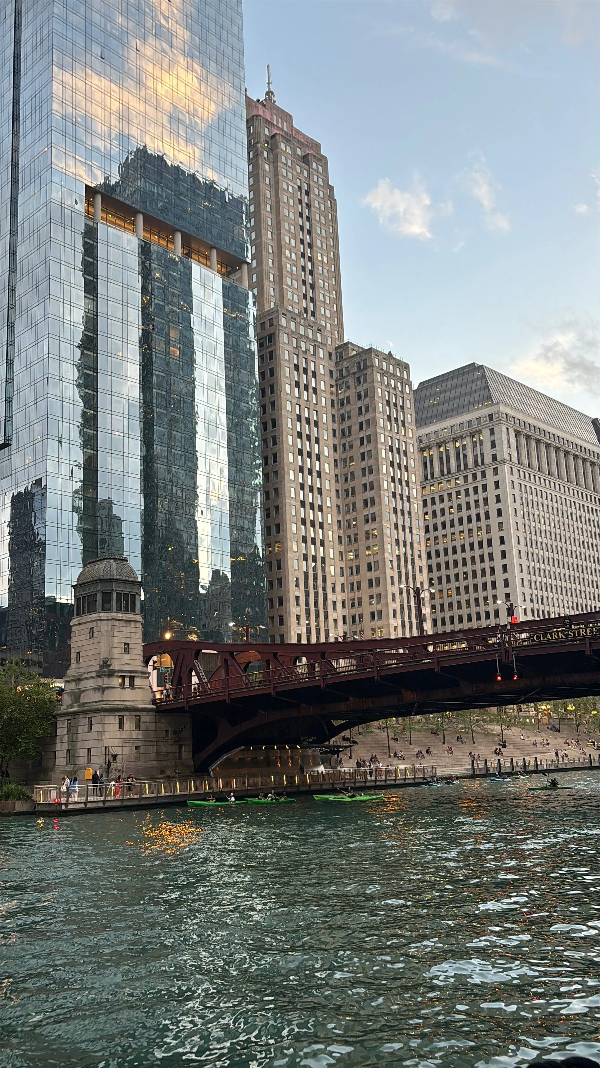 Cityscape with tall buildings, a bridge over water, and kayakers and pedestrians along the water's edge in the foreground.