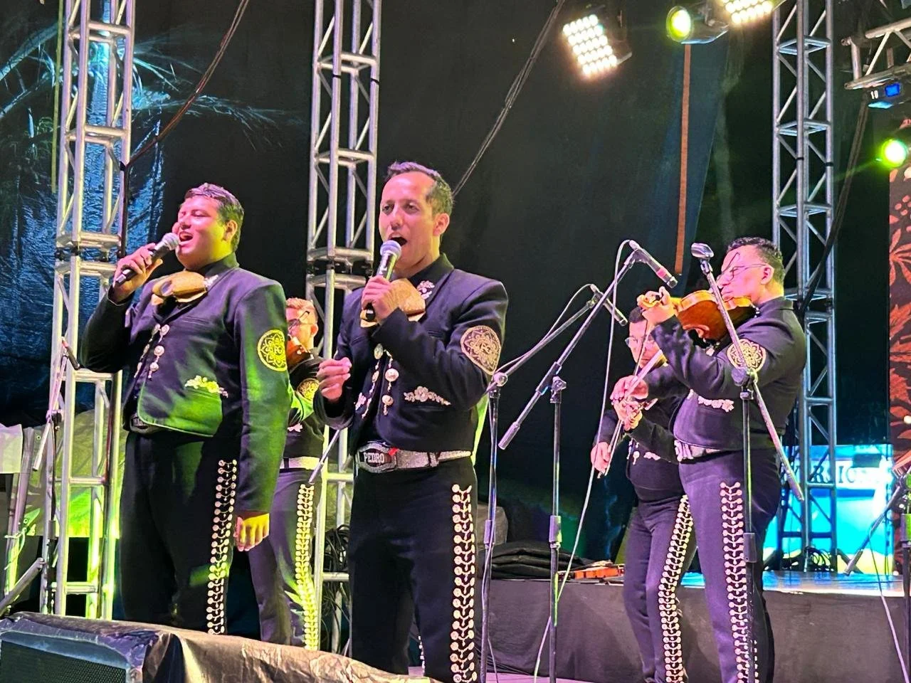 Grupo de músicos mexicanos vestidos con trajes tradicionales de mariachi tocando instrumentos en un escenario con luces y estructura metálica.
