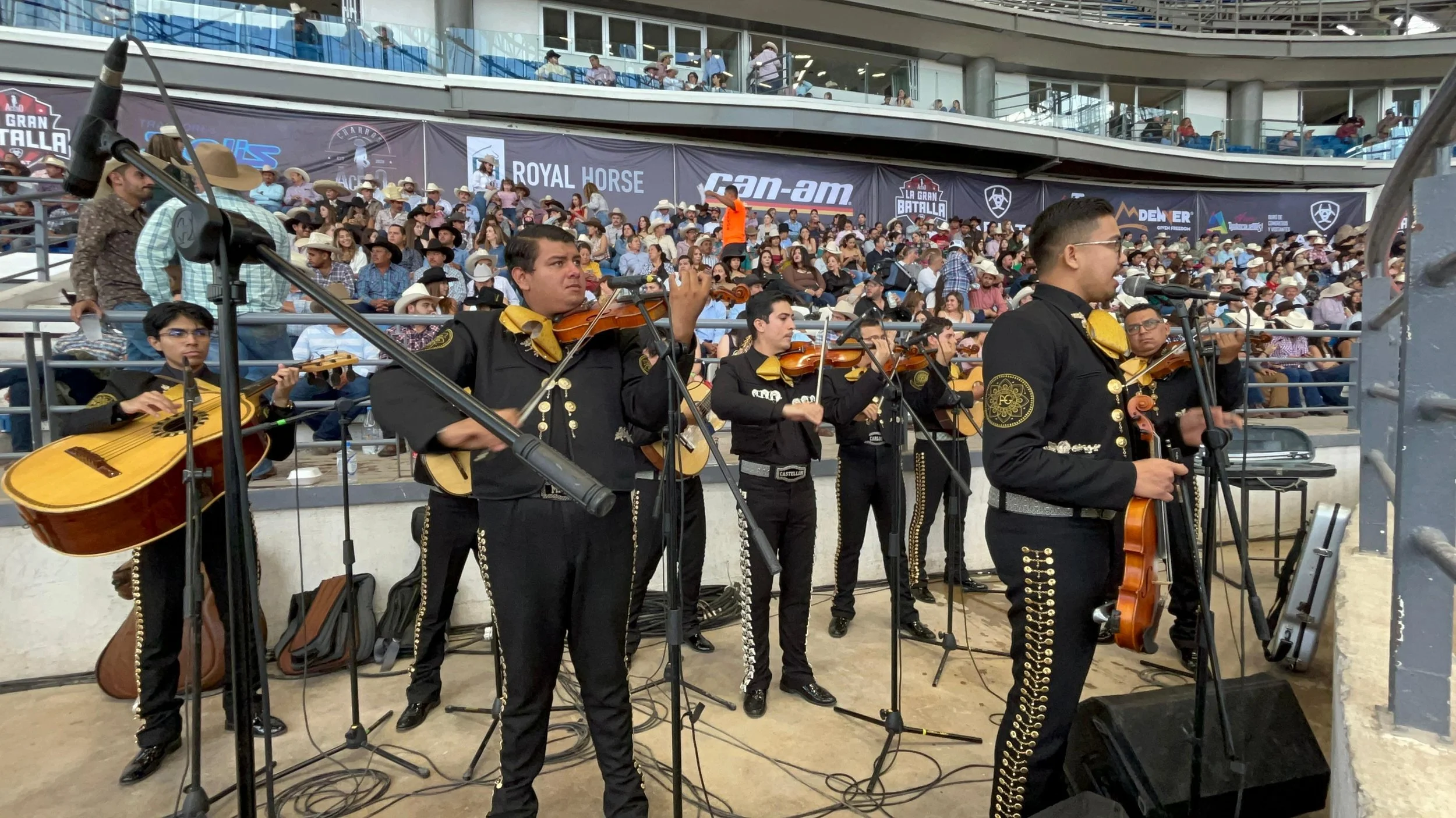 Orquesta de mariachi tocando en un escenario durante un evento con un público grande en las gradas.