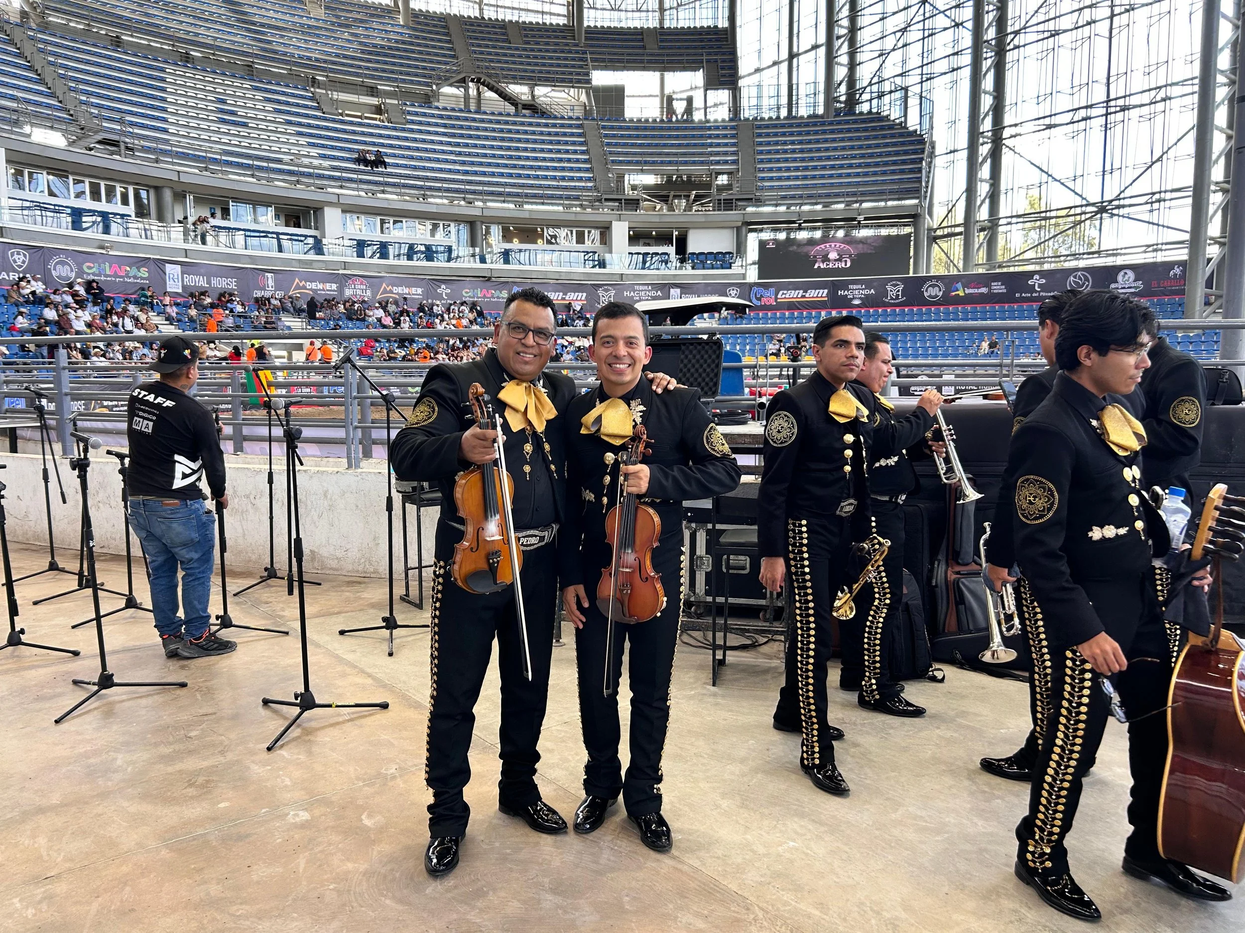Cuatro músicos de mariachi vestidos con trajes negros y lazos dorados, algunos con instrumentos, en un espectáculo en un estádio con público.