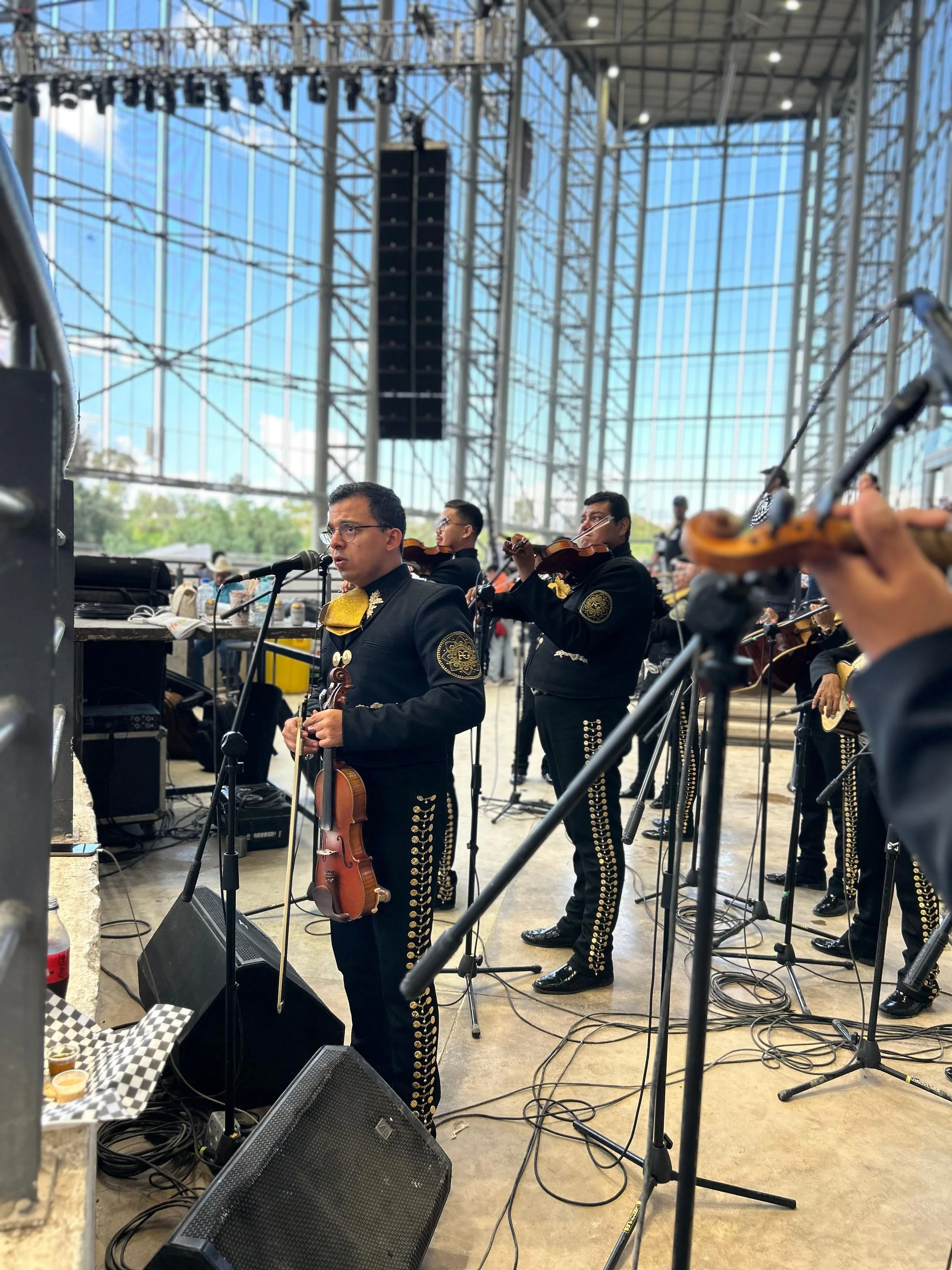 Una orquesta tradicional mexicana tocando instrumentos en un escenario amplio y techado, con músicos vestidos con traje de mariachi.
