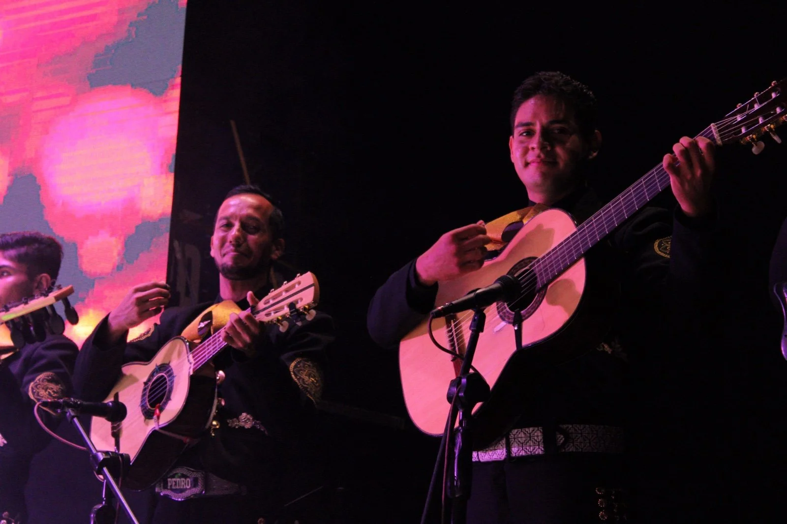 Dos músicos tocando guitarras en un escenario con fondo de pantalla colorido y oscuro.