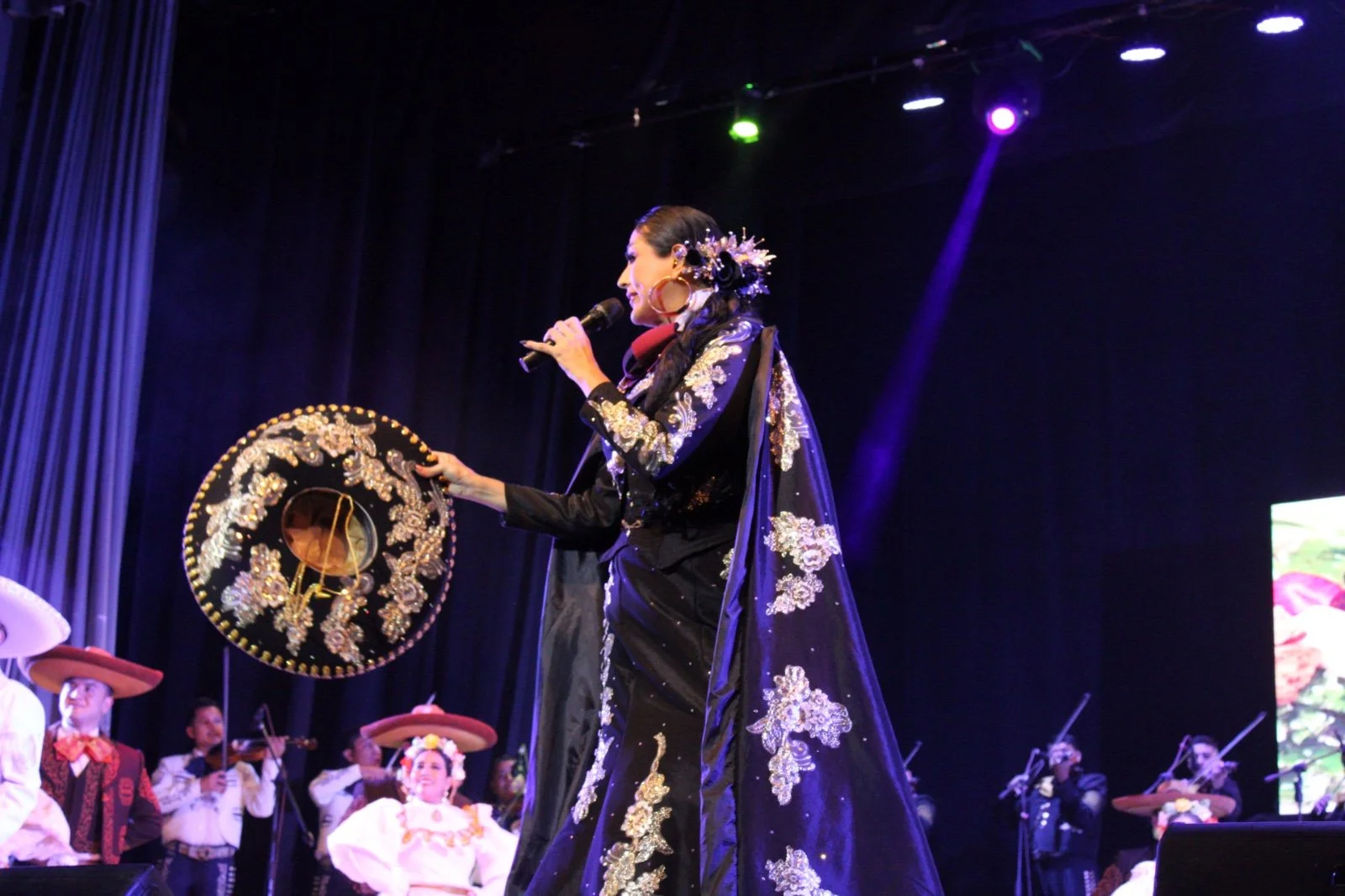 Una mujer vestida con traje tradicional mexicano en un escenario, acompañada de un grupo de personas con trajes típicos, en un espectáculo cultural con luces de colores.