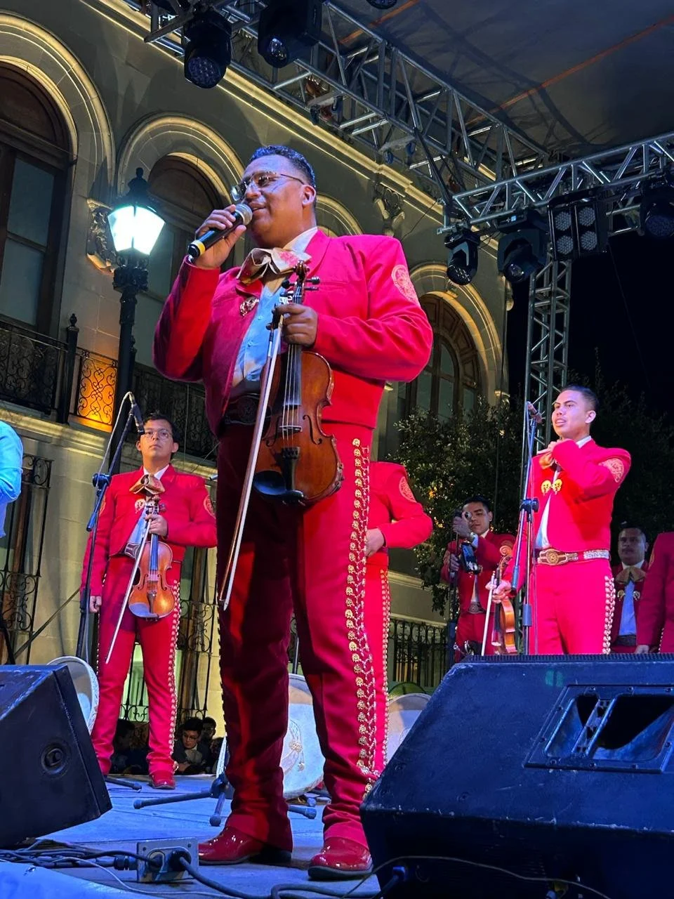 Un grupo de músicos vestidos con trajes tradicionales de mariachi en un escenario al aire libre, tocando instrumentos como violines y cantando.