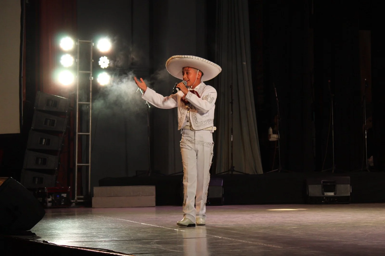 Niña vestida con traje típico mexicano, sosteniendo un micrófono en un escenario, con luces brillantes detrás.