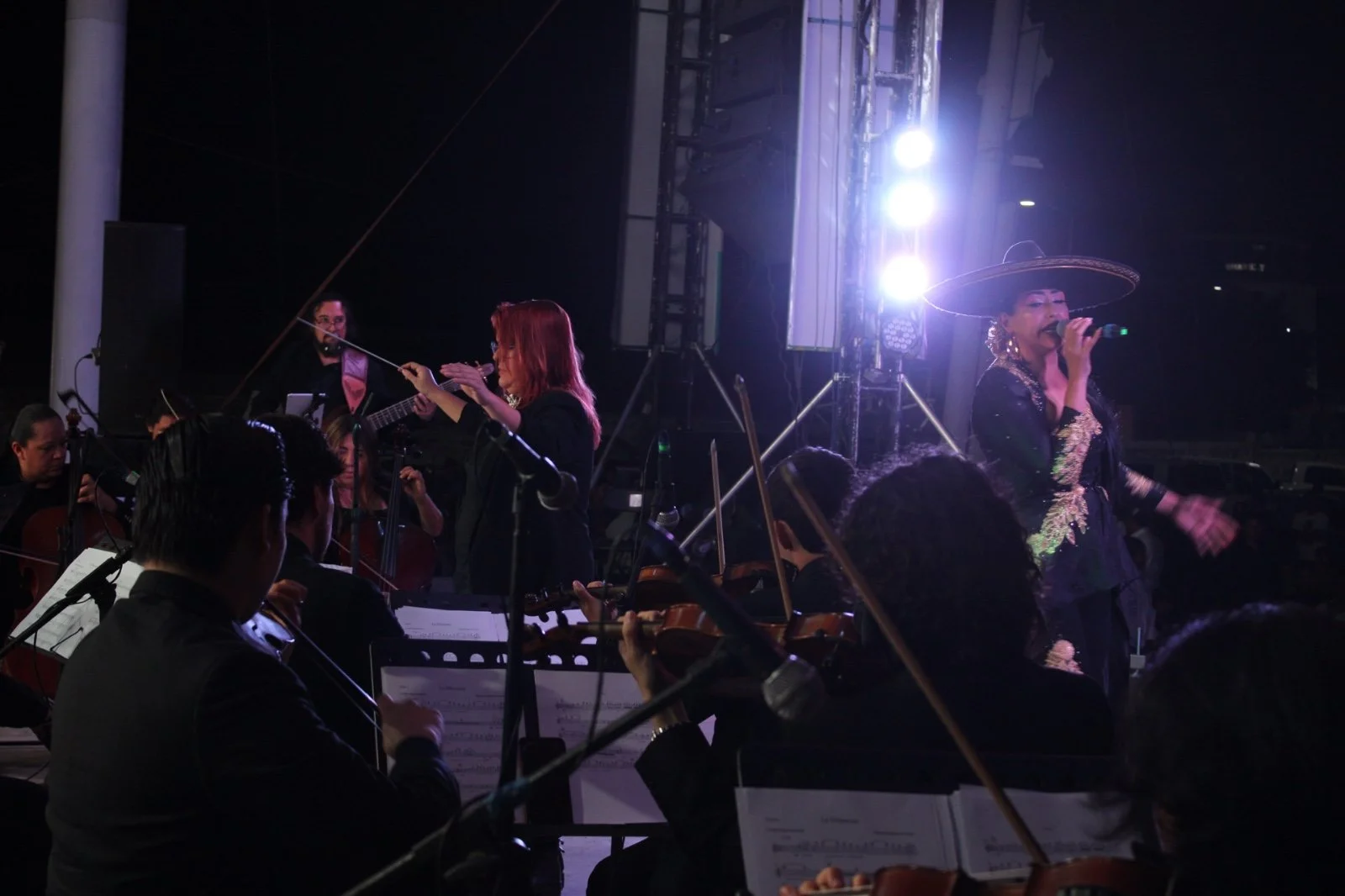 Una orquesta en un escenario durante un concierto, con una cantante con sombrero grande y vestimenta tradicional mexicana, y músicos con instrumentos de cuerda y guitarra en un entorno oscuro con iluminación fuerte.