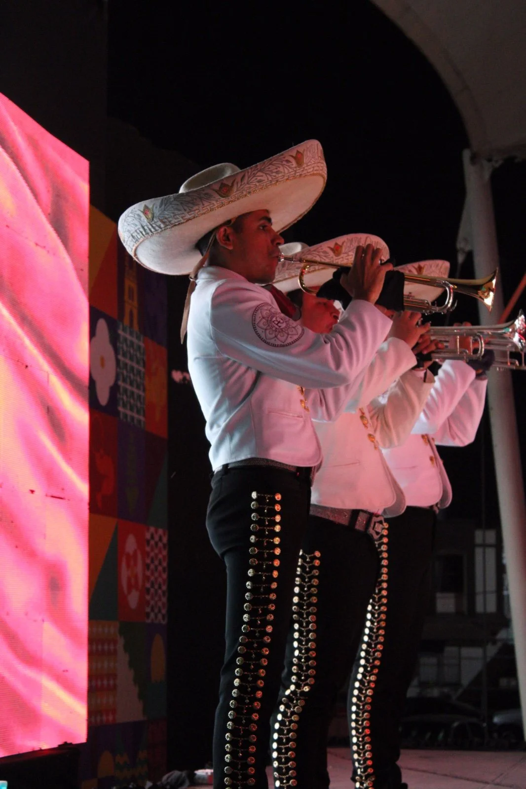 Tres músicos tocando trompetas en un escenario, vestidos con trajes tradicionales y sombreros grandes.