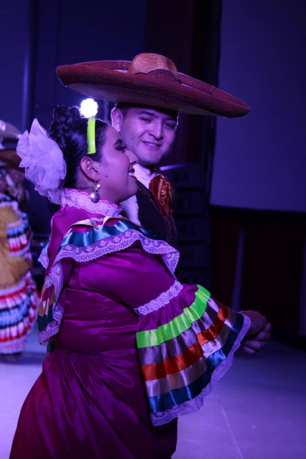 Pareja vestida con trajes tradicionales mexicanos, ella con un vestido de colores vibrantes y él con un sombrero grande, participando en una danza típica.